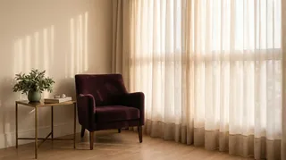 Poltrona de leitura vazia ao lado de uma janela alta com cortina de linho, luz de fim de tarde